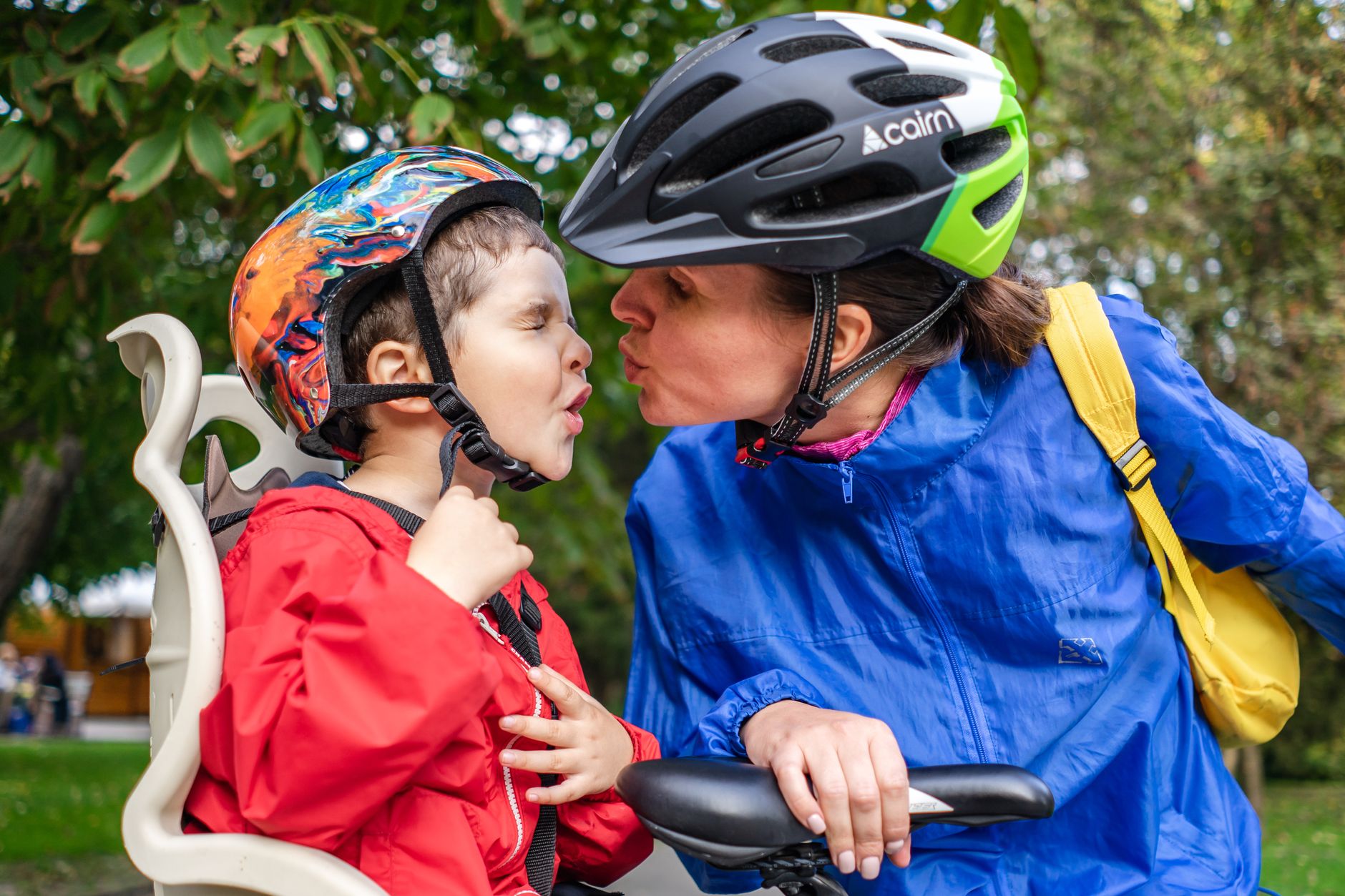 A mother and son share a kiss while wearing safety helmets on a bike ride outdoors.