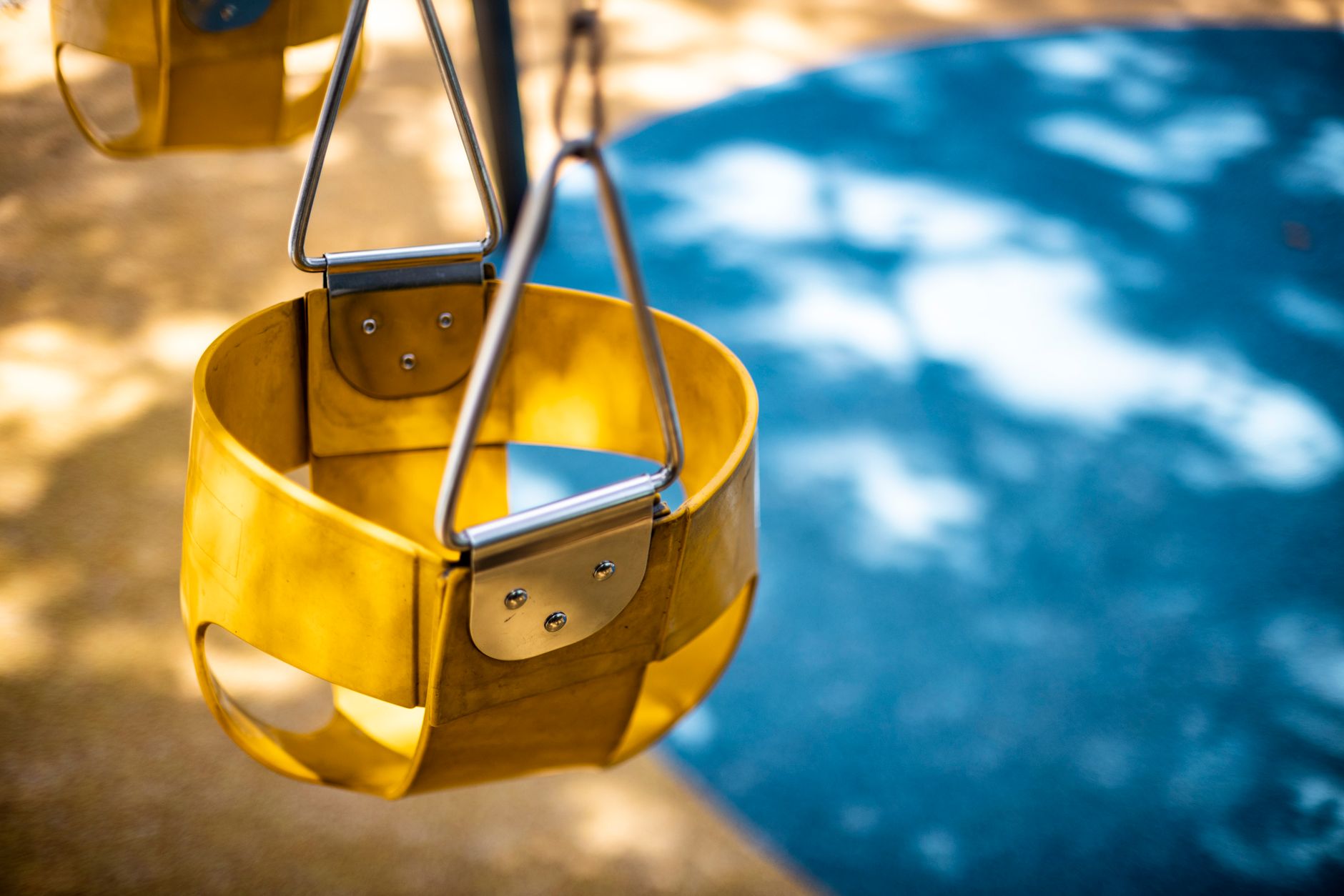 Close-up of a yellow playground swing seat in a sunlit park setting, casting shadows on the pavement.