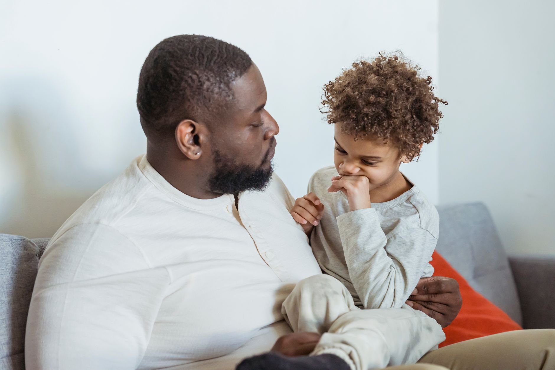 Calm African American father looking at cute black son sitting on knees on couch near wall in light room at home