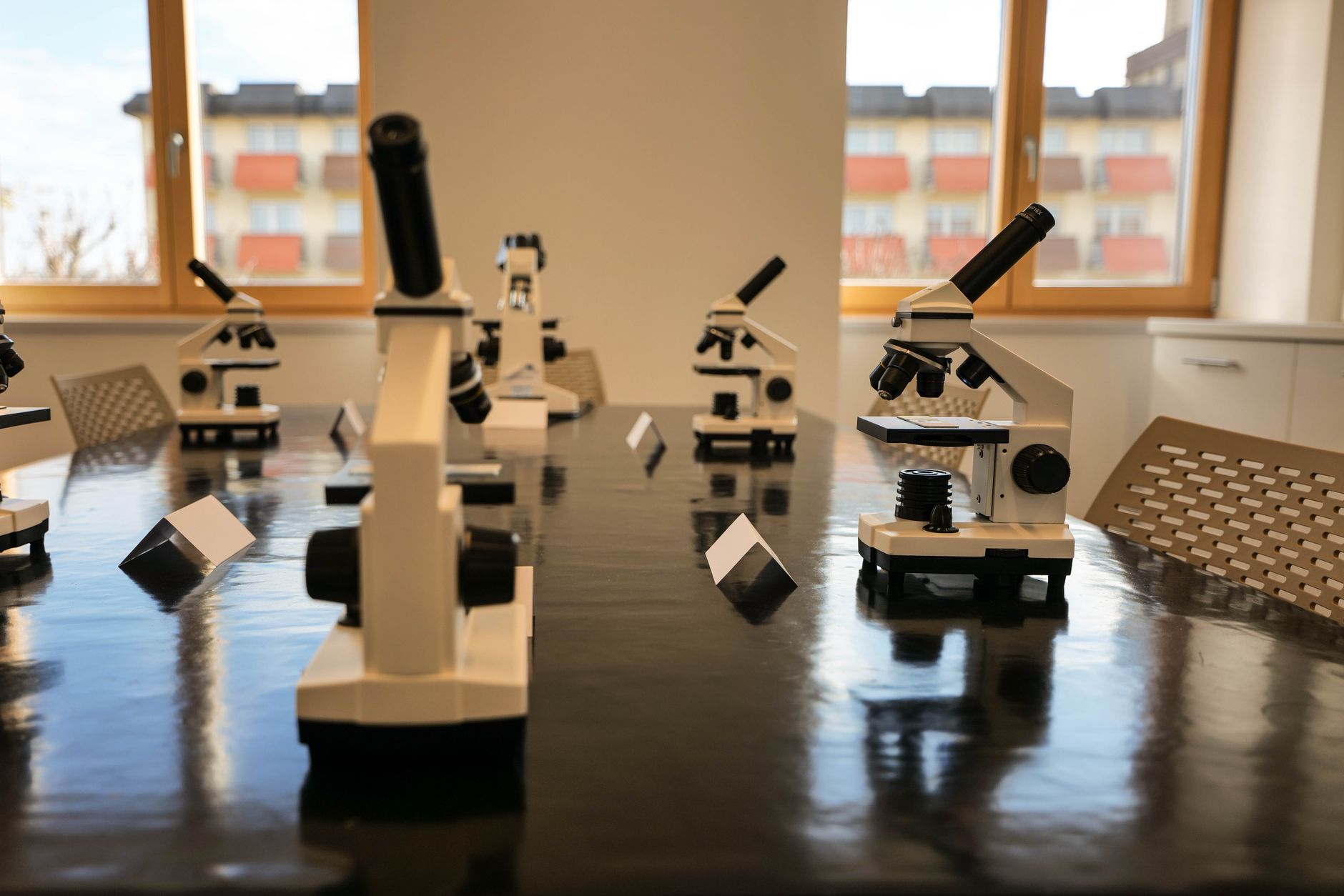 A modern laboratory classroom featuring microscopes on a glossy table, with large windows and natural light.