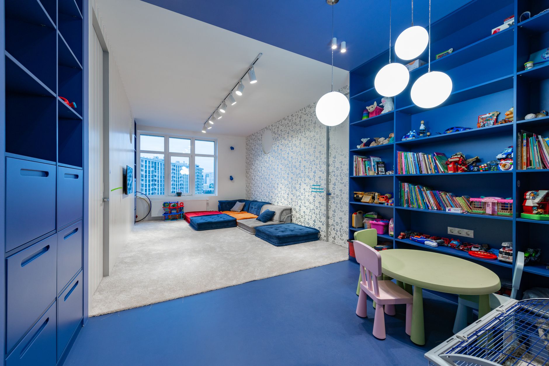 Interior of playroom for children with blue shelves with books and toys placed near light room with colorful mattresses in daytime