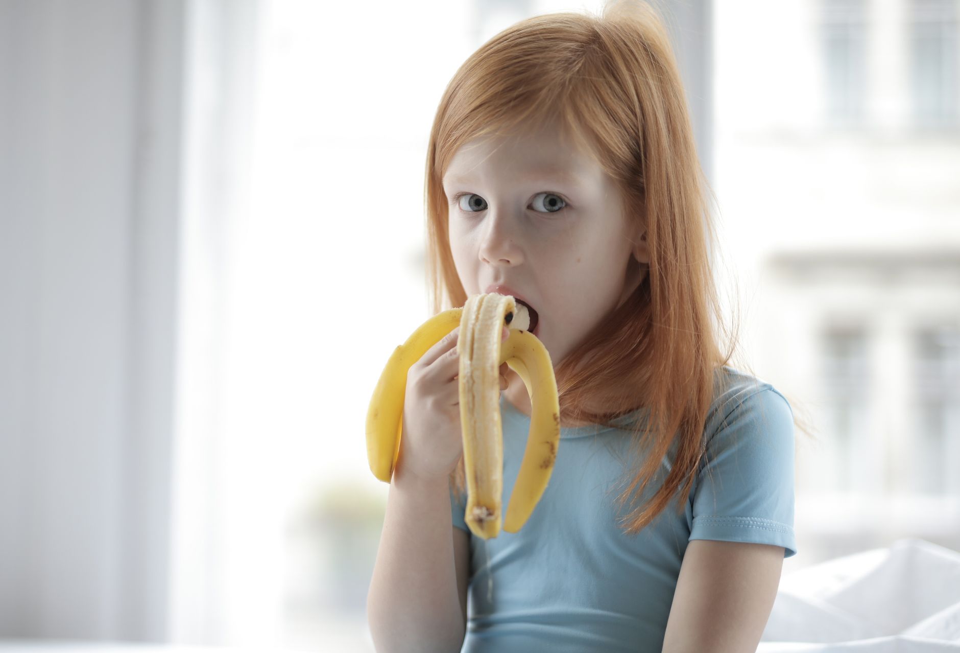 Portrait of a girl eating a banana indoors, emphasizing child nutrition and healthy eating.