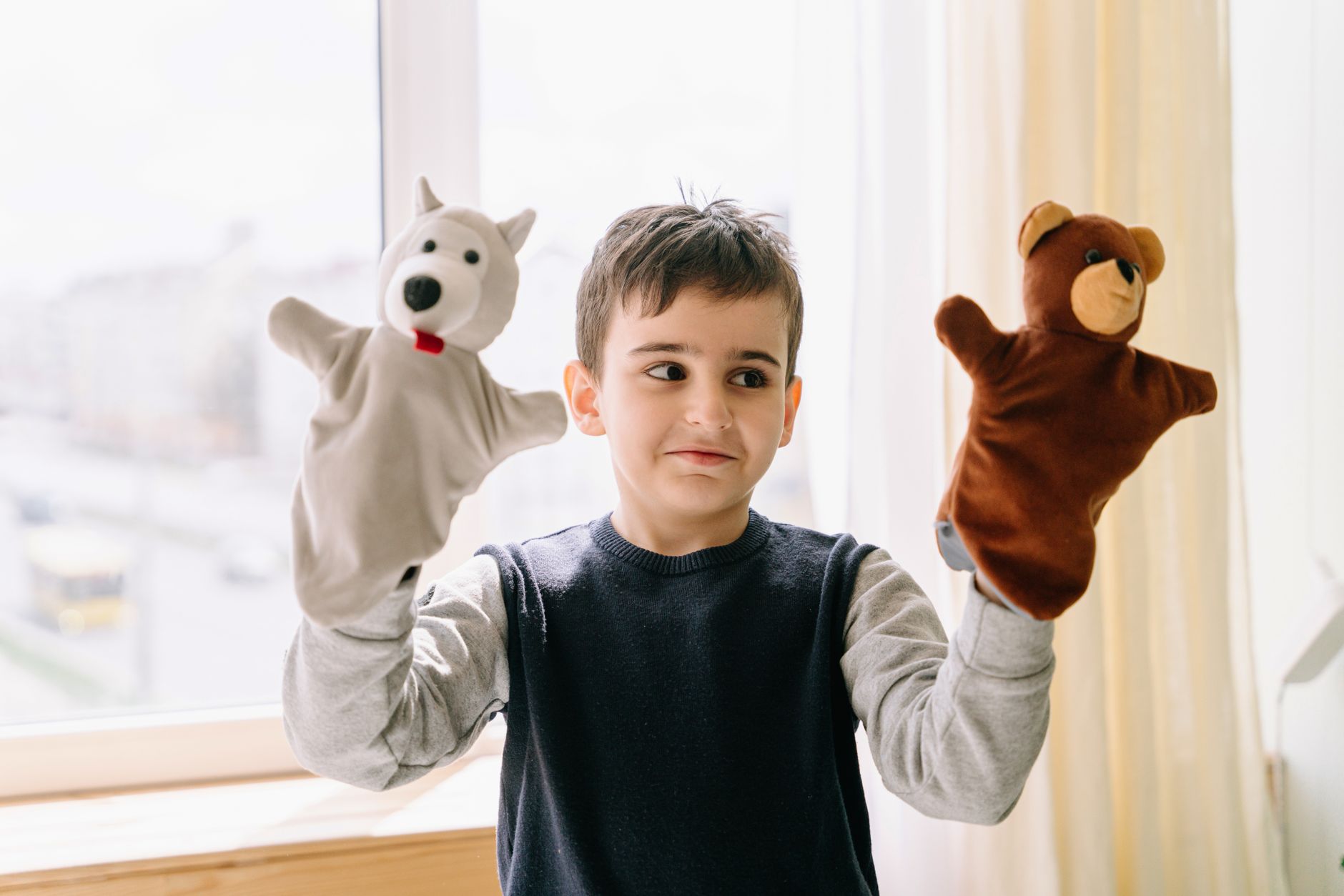 A young boy joyfully plays with bear and polar bear hand puppets indoors. Bright daylight setting.